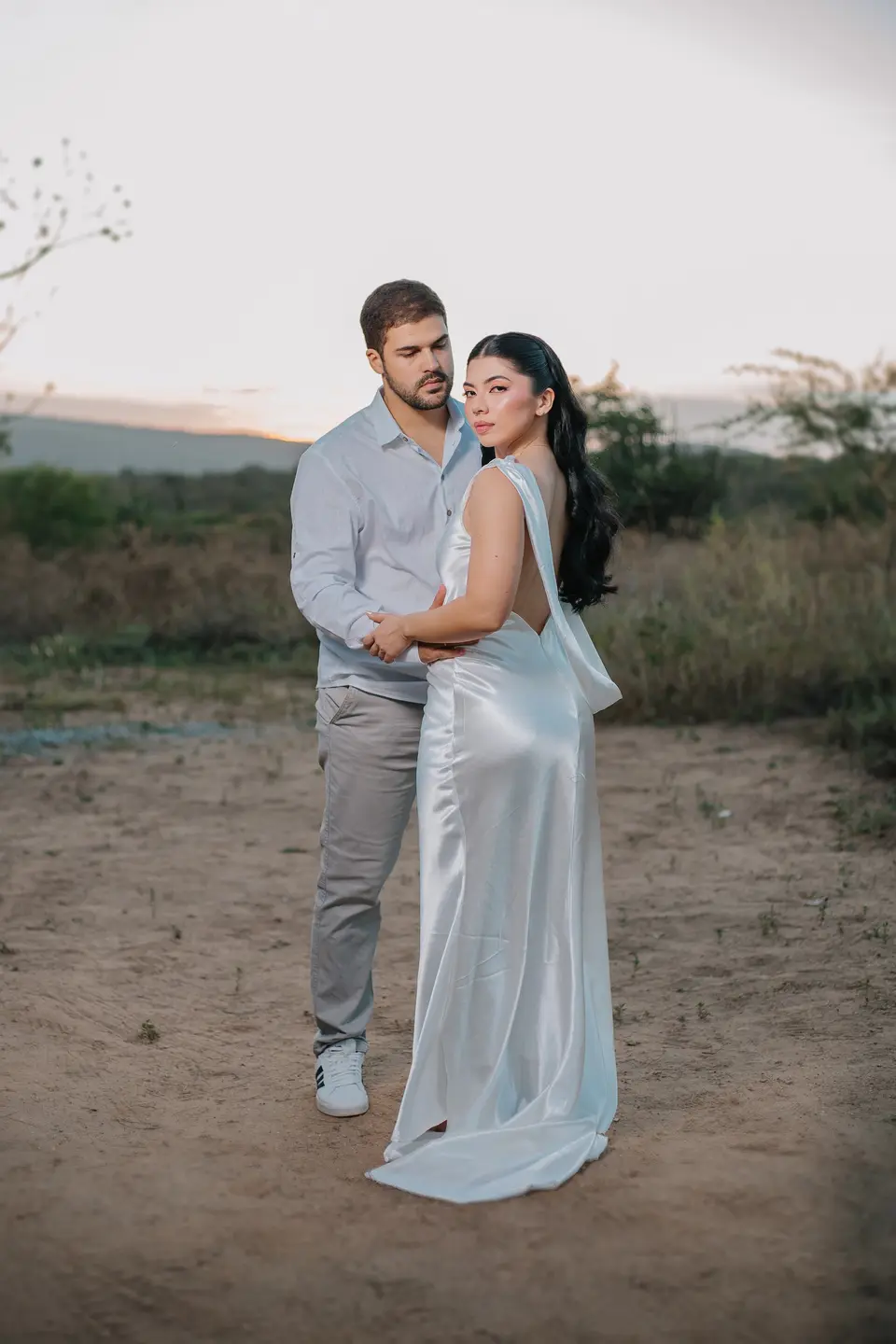 Jessica e Felipe abraçados em um campo ao entardecer; ela olha para a câmera por cima do ombro